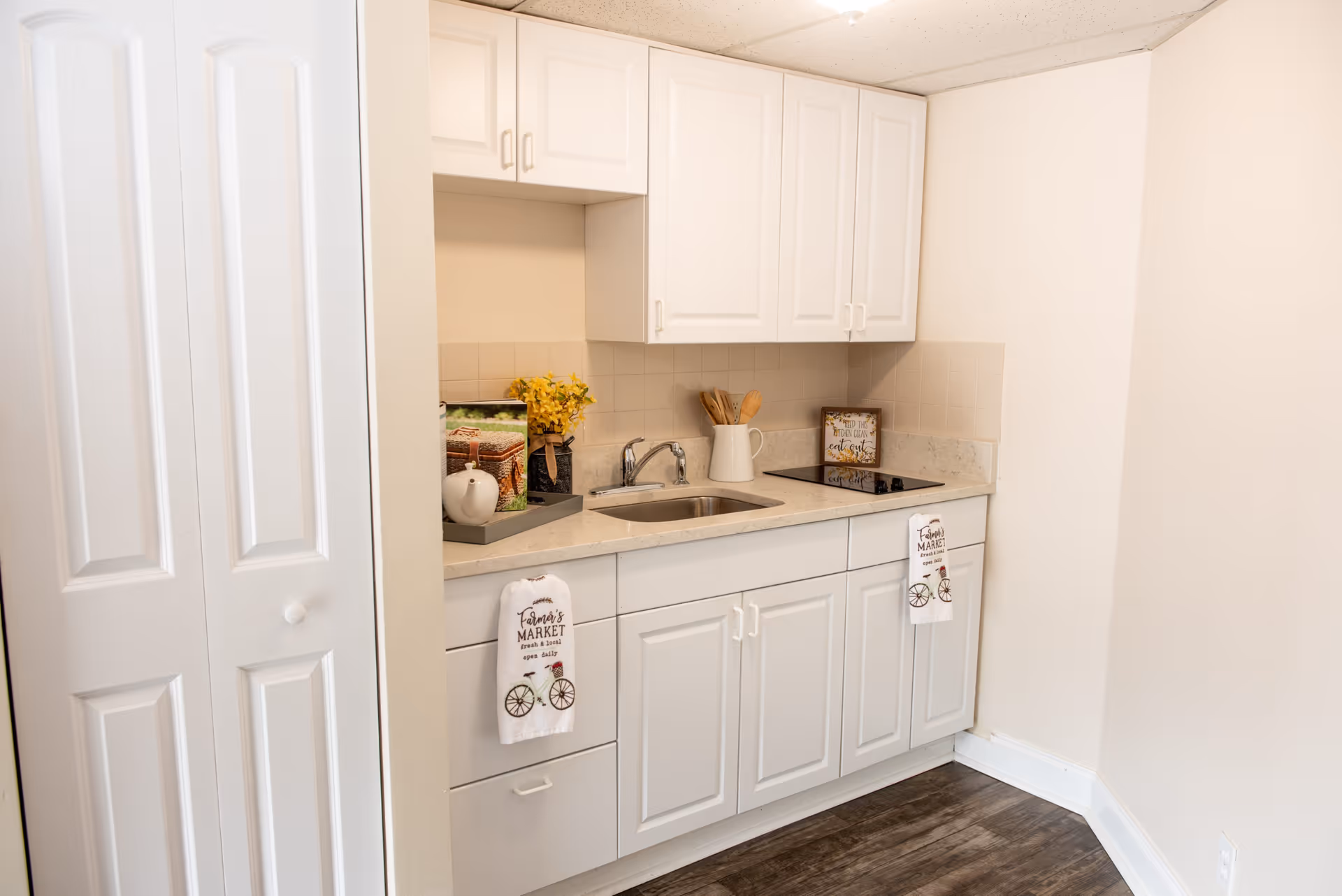 A small kitchen area with white cabinets, a countertop with a sink, a stovetop, and decorative items including a tray with a teapot and flowers, wooden utensils in a white pitcher, and a framed sign. Two white towels with a bicycle and 'Farmers Market' text hang from the cabinet handles. The floor is dark wood and the walls are light-colored.