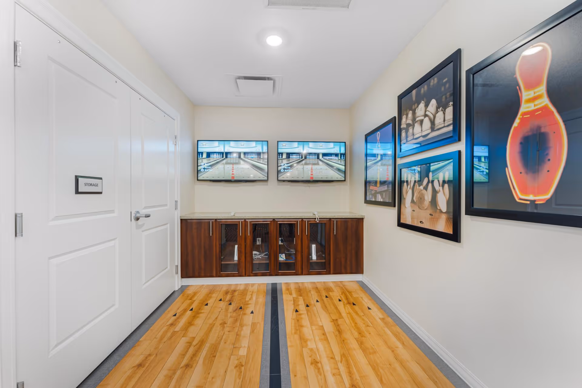 A small room with light-colored walls and wooden flooring featuring a black stripe down the center. On the left side, there are two white doors, one labeled 'STORAGE'. On the far wall, there is a wooden cabinet with glass doors and a countertop above it. Above the cabinet, two flat-screen TVs display images of a bowling alley. The right wall is decorated with framed pictures of bowling pins and bowling scenes.
