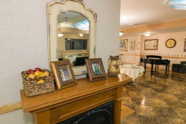 Wooden mantel with a mirror, framed photos and a basket of fruit in the foreground overlooking a communal dining/activity room with tables and a piano.