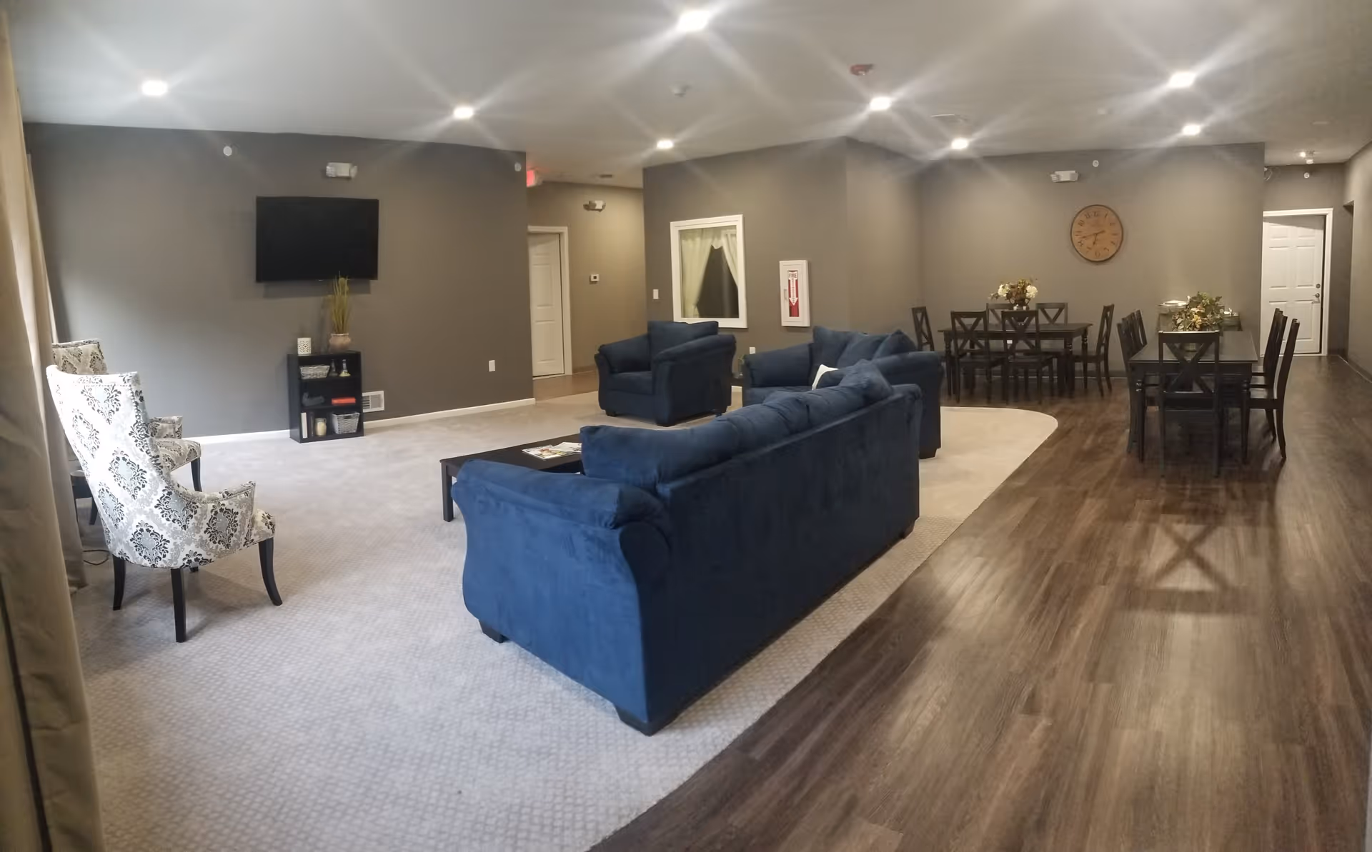 A spacious living room area with blue sofas and patterned armchairs on a light carpet, a wall-mounted TV, and a small black shelf with decorative items. Adjacent to the living area is a dining space with two dark wood dining tables and chairs, a wall clock, and floral centerpieces. The room has gray walls, recessed ceiling lights, and a wood floor in the dining area.