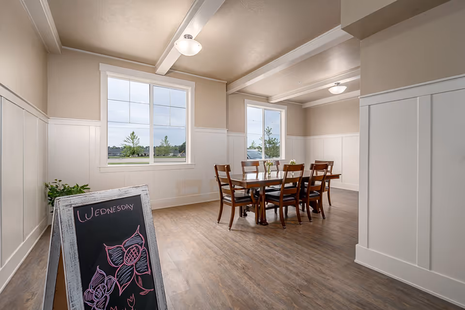 A bright dining room with wooden flooring and beige walls featuring white wainscoting. There is a wooden dining table with six chairs in the center of the room, and two large windows letting in natural light. A chalkboard with floral drawings and the word 'Wednesday' written on it is placed near the entrance.