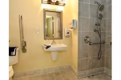 Accessible bathroom with a wall-mounted sink, a mirror above it, a soap dispenser, a grab bar next to the sink, and a tiled walk-in shower with a handheld showerhead and grab bars.