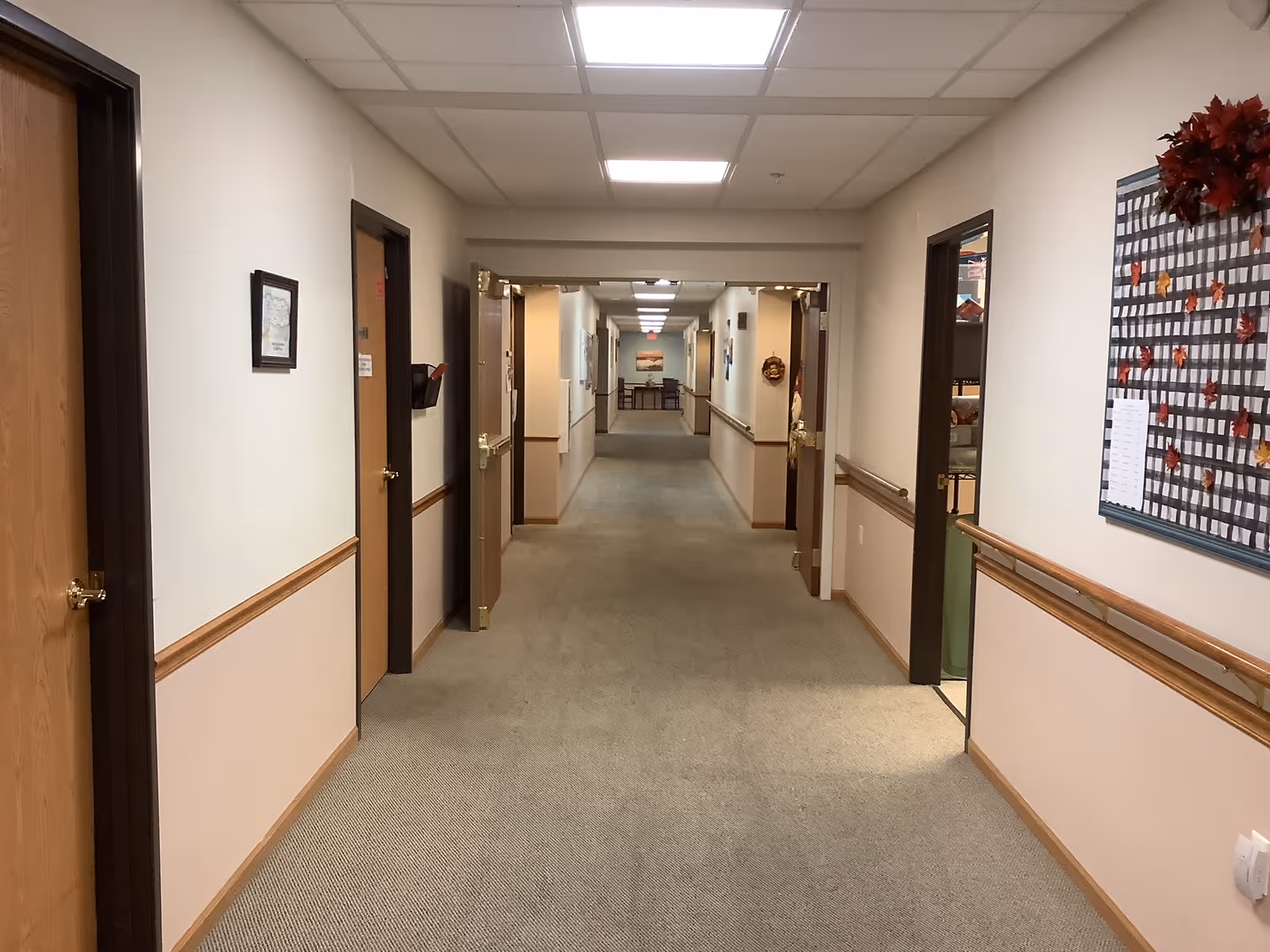 Carpeted interior hallway in a senior living facility with doors and handrails on both sides and overhead lighting.