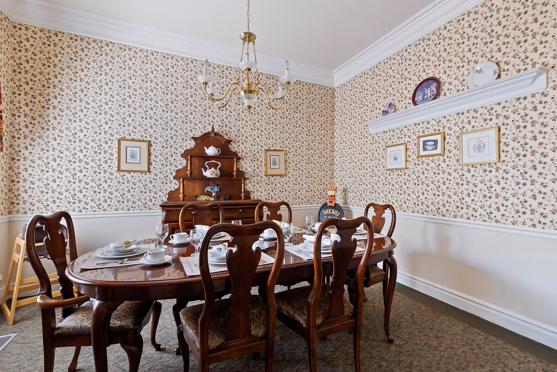A traditional dining room with a wooden dining table set for six people with white plates, cups, and glasses. The room features floral wallpaper, a wooden hutch with decorative items, framed pictures on the walls, and a chandelier hanging from the ceiling.