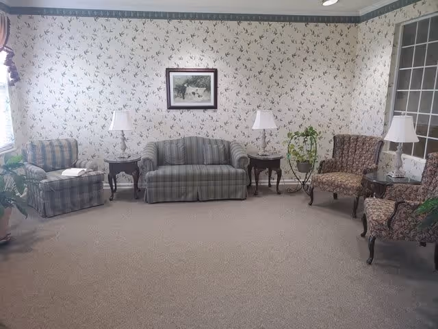 A sitting area in an assisted living facility with a plaid sofa and armchair, two floral patterned armchairs, three side tables each with a lamp, a framed picture on floral wallpaper, and a potted plant near a large window.