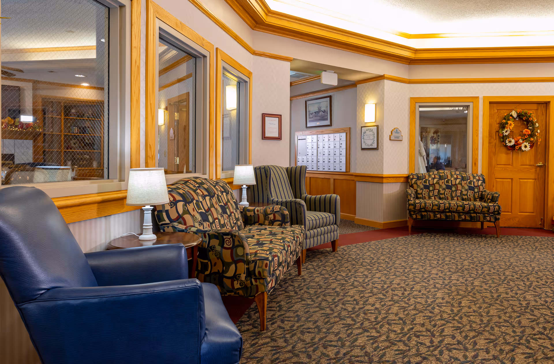 A cozy seating area in an assisted living facility with patterned armchairs and a blue leather chair arranged along the wall. There are two small lamps on side tables between the chairs. The walls have wood trim and there are windows with wire mesh glass. A door decorated with a fall-themed wreath is visible, along with a set of mailboxes on the wall.