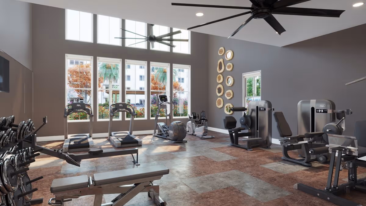A spacious fitness room with large windows letting in natural light. The room contains various exercise equipment including treadmills, stationary bikes, weight machines, and a bench with free weights. The walls are painted gray and decorated with circular wooden art pieces. Two large ceiling fans are mounted on the ceiling.