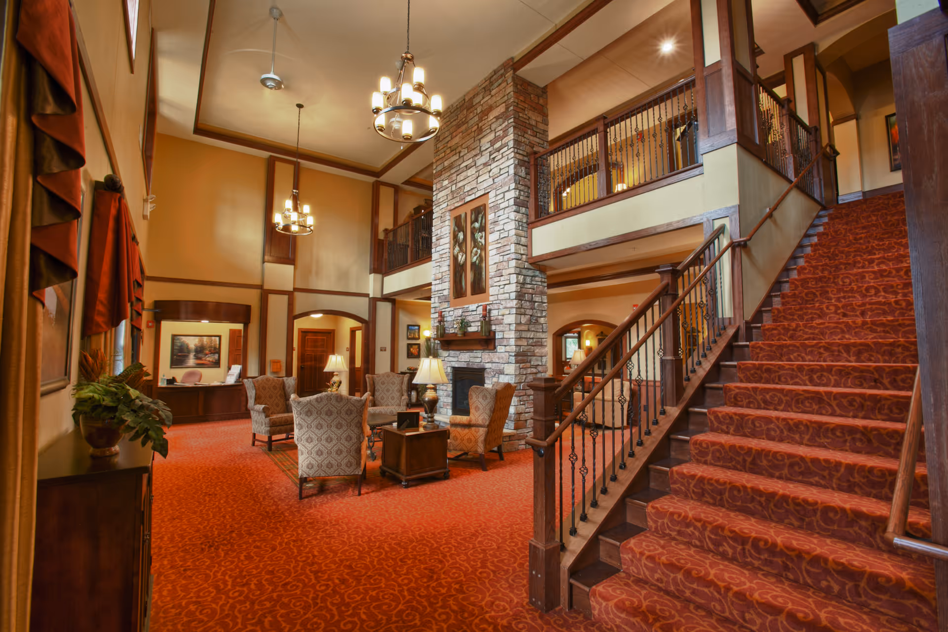 Spacious two-story lounge with red carpet, seating around a stone fireplace and a carpeted staircase leading to an upper balcony.