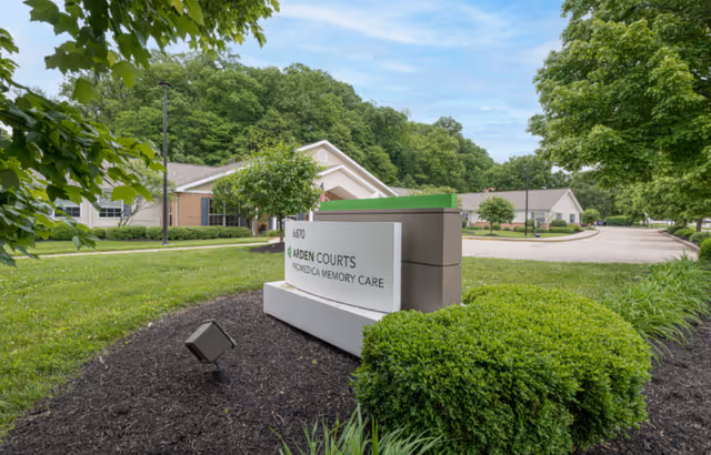 Entrance sign reading "Arden Courts ProMedica Memory Care" in landscaped grounds in front of a single-story memory care building surrounded by trees.
