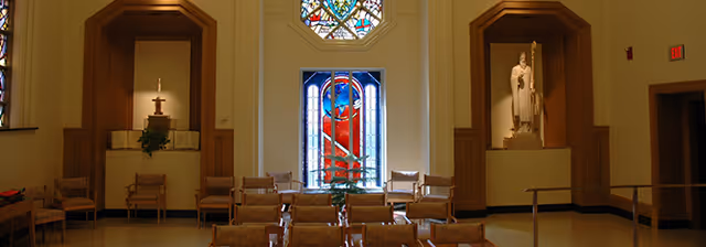 Interior of a small chapel with rows of chairs facing a central stained-glass window flanked by alcoves containing religious statues and altars.