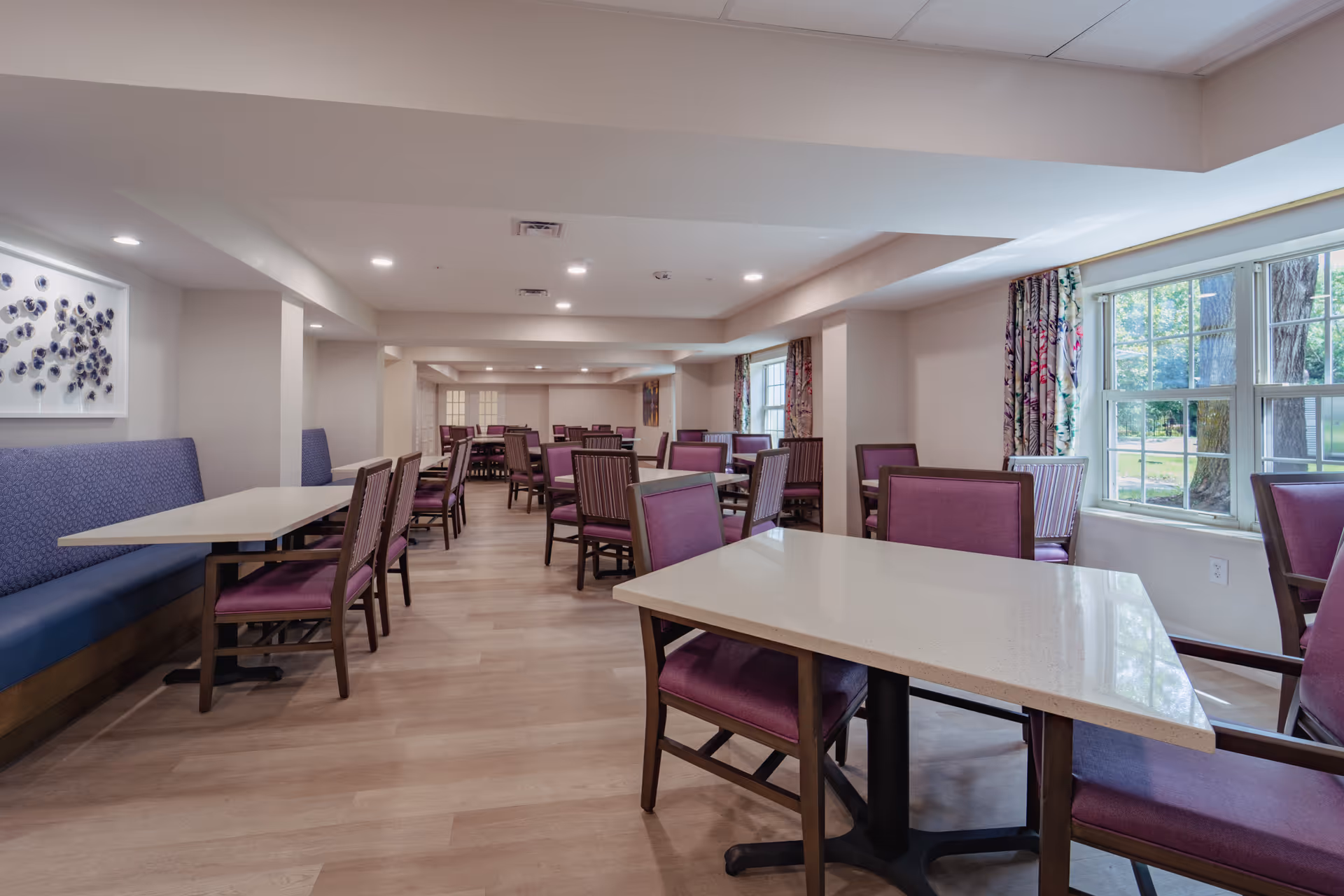 A spacious dining room with multiple tables and chairs featuring purple upholstery. The room has large windows with floral curtains allowing natural light to enter. The walls are light-colored, and there is a piece of abstract art on one wall. The floor is wooden, and the ceiling has recessed lighting.