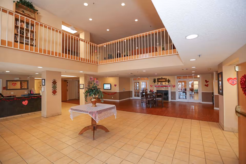 Spacious two-story senior living facility lobby with a central table and floral arrangement, reception desk, and seating area.