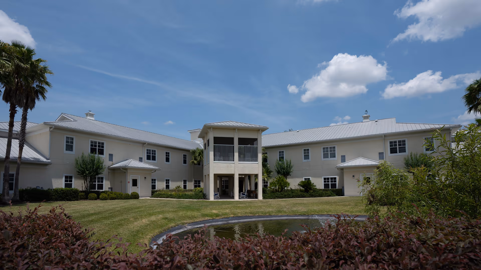 Exterior view of a two-story senior living facility building with a light-colored facade and metal roof under a blue sky with scattered clouds. The building features a central covered balcony area supported by columns, surrounded by well-maintained green lawns, palm trees, bushes, and a small pond with a fountain in the foreground.