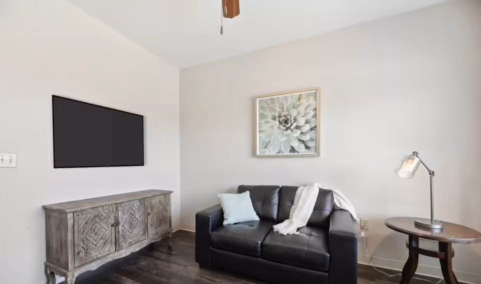 A small living room with a black leather loveseat adorned with a light blue pillow and a white throw blanket. Above the loveseat hangs a framed floral artwork. To the right is a round wooden side table with a modern lamp. On the left wall, a flat-screen TV is mounted above a decorative wooden cabinet. The room has light-colored walls and dark wood flooring.