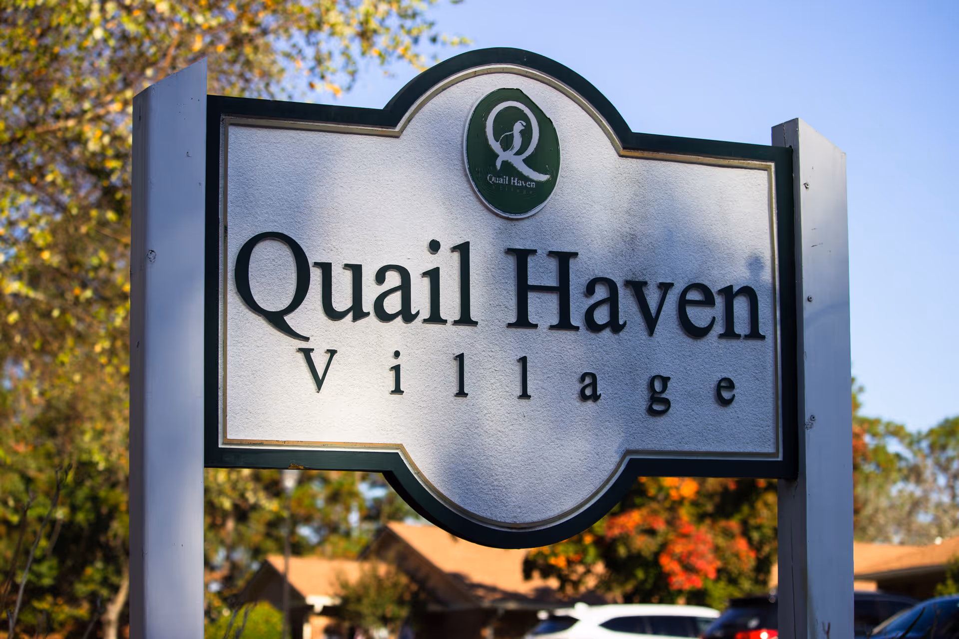 A close-up view of a sign for Quail Haven Village, an outdoor scene with trees and houses in the background under a clear blue sky.
