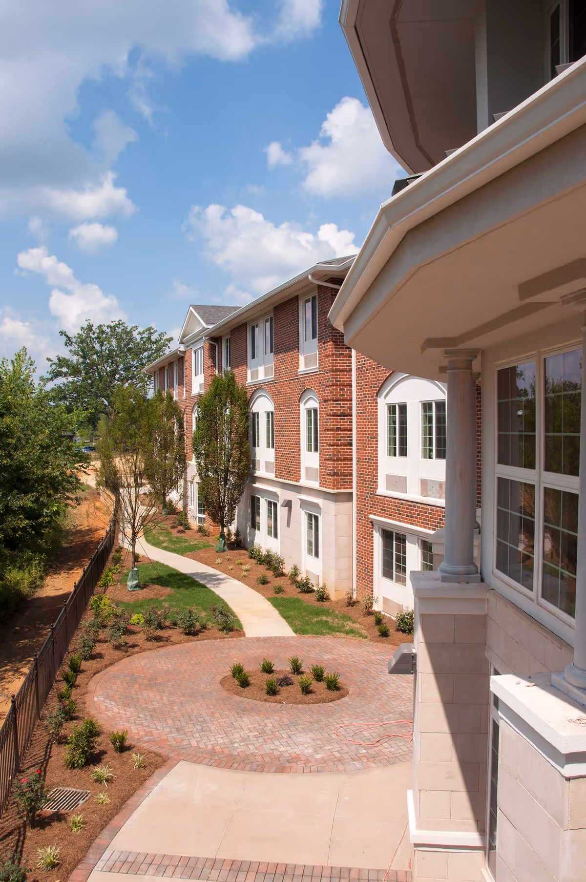 Exterior view of a multi-story brick building with white trim, surrounded by a landscaped garden with young trees and shrubs. A curved paved walkway and circular brick patio are visible under a partly cloudy blue sky.