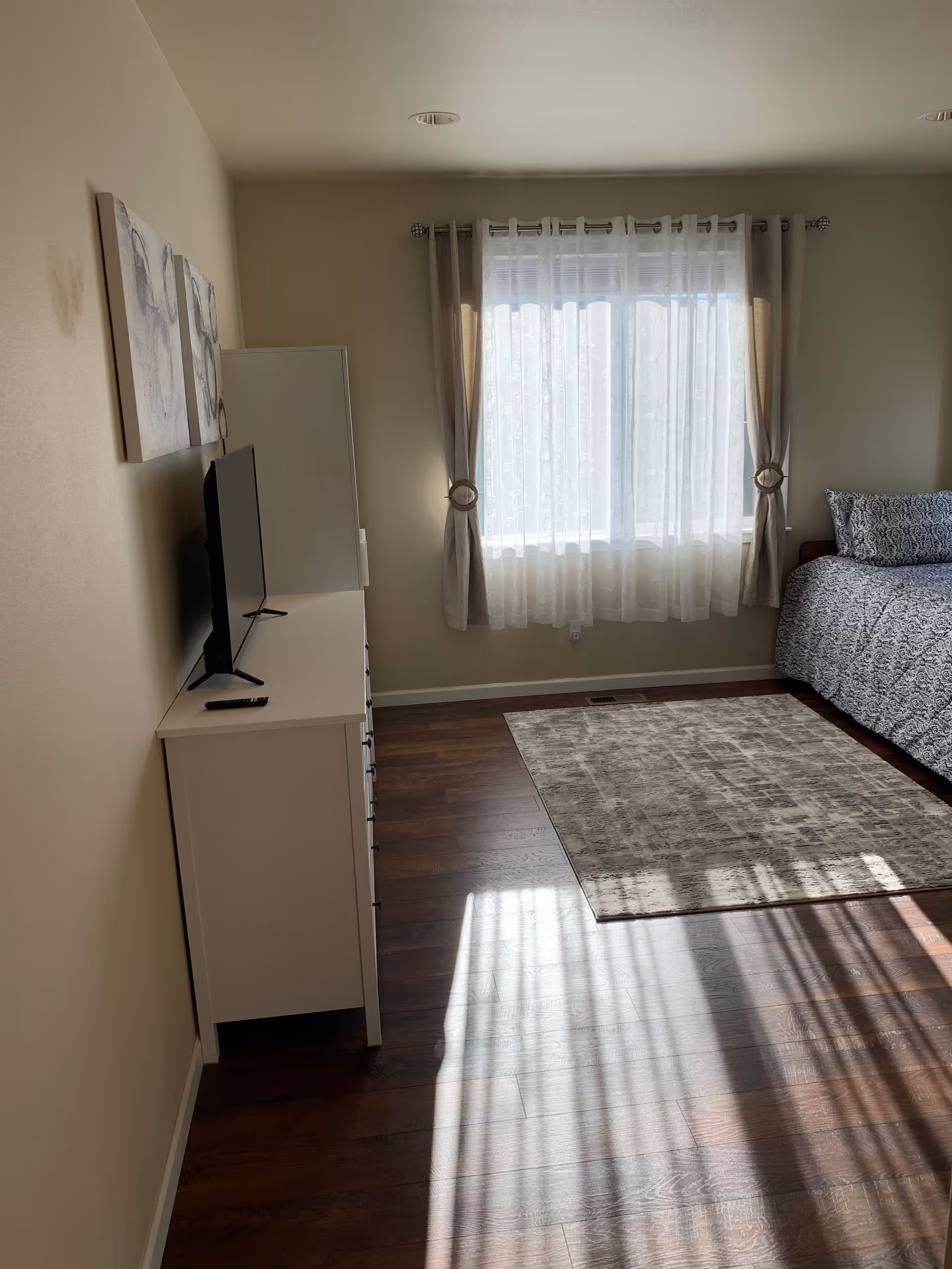 Sunlit bedroom with a bed, dresser topped by a TV, a rug on hardwood floor, and a window with sheer curtains.