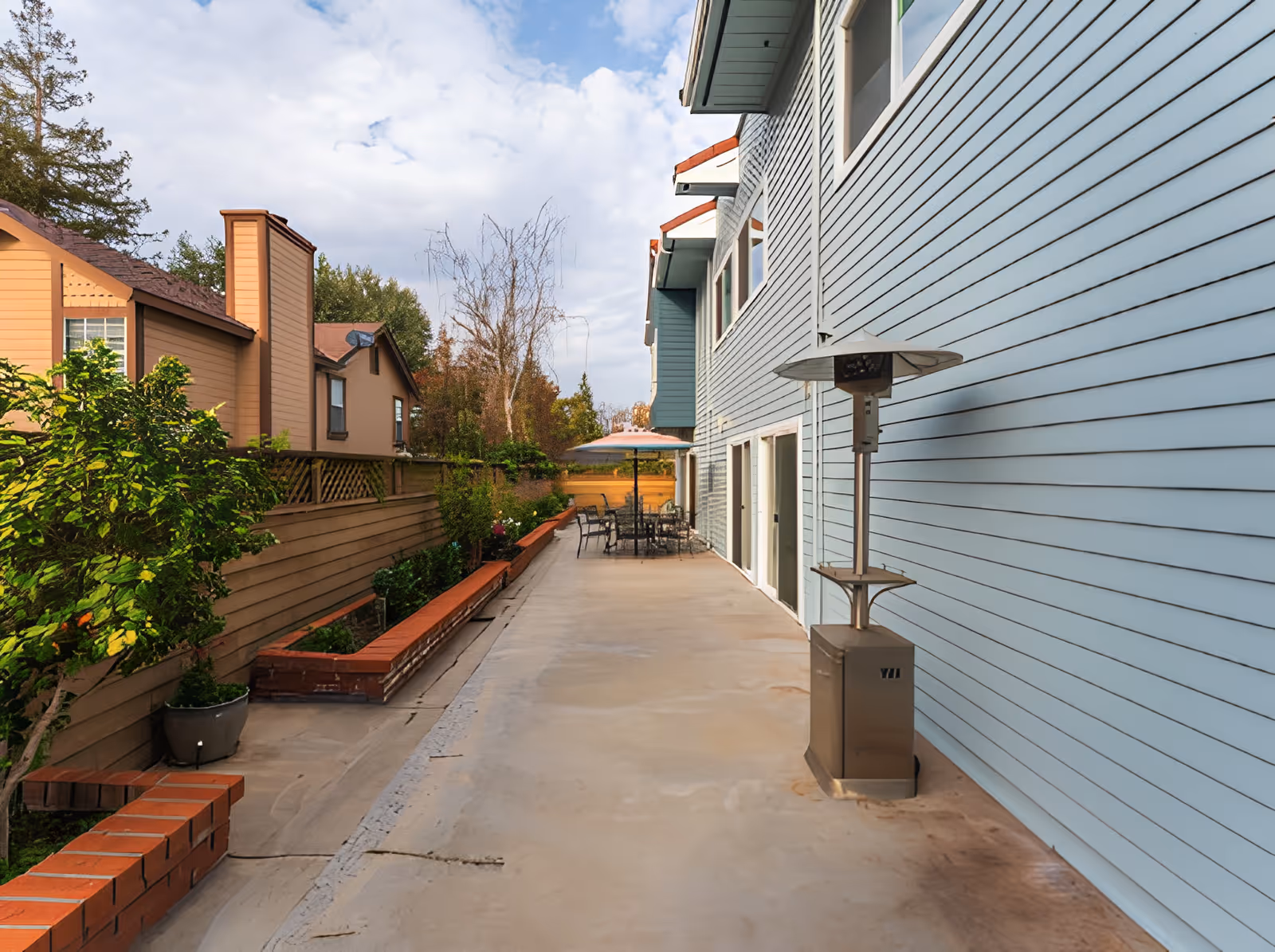 Outdoor patio area beside a light blue building with several windows. The patio has a concrete floor, a tall outdoor heater, a table with chairs, and an umbrella. There is a wooden fence with plants and trees on the left side, and neighboring houses are visible in the background under a partly cloudy sky.