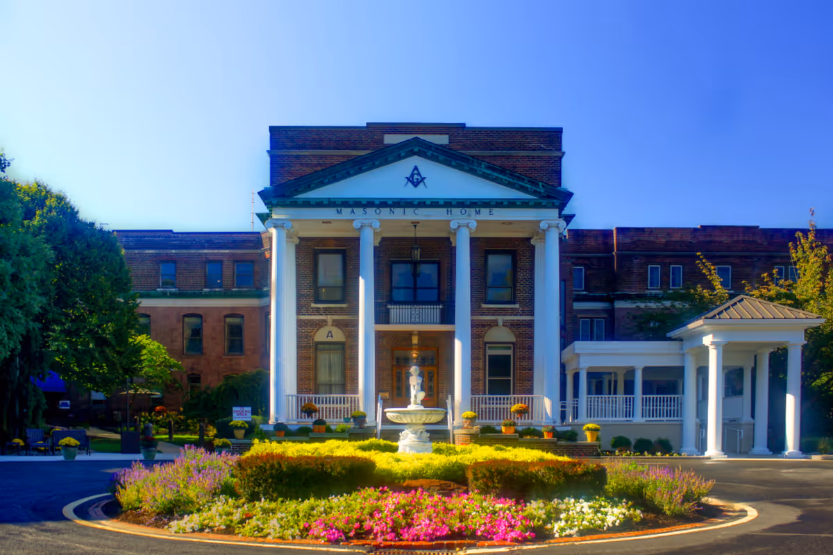 Front exterior view of a large brick building with white columns and a triangular pediment displaying a Masonic symbol. The building is labeled 'Masonic Home' and is surrounded by a circular driveway with a central fountain and colorful flower beds. Trees and greenery are visible on either side under a clear blue sky.