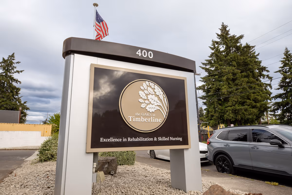 Outdoor sign for The Oaks at Timberline displaying the facility's name and the phrase 'Excellence in Rehabilitation & Skilled Nursing' with an American flag on top, surrounded by trees, parked cars, and a cloudy sky.
