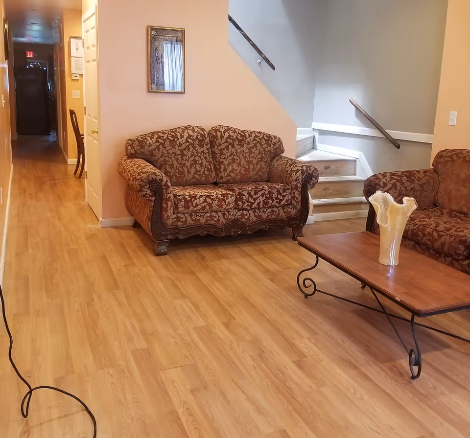 A living room with patterned sofas, a wooden coffee table topped by a vase, and a staircase and hallway in the background.