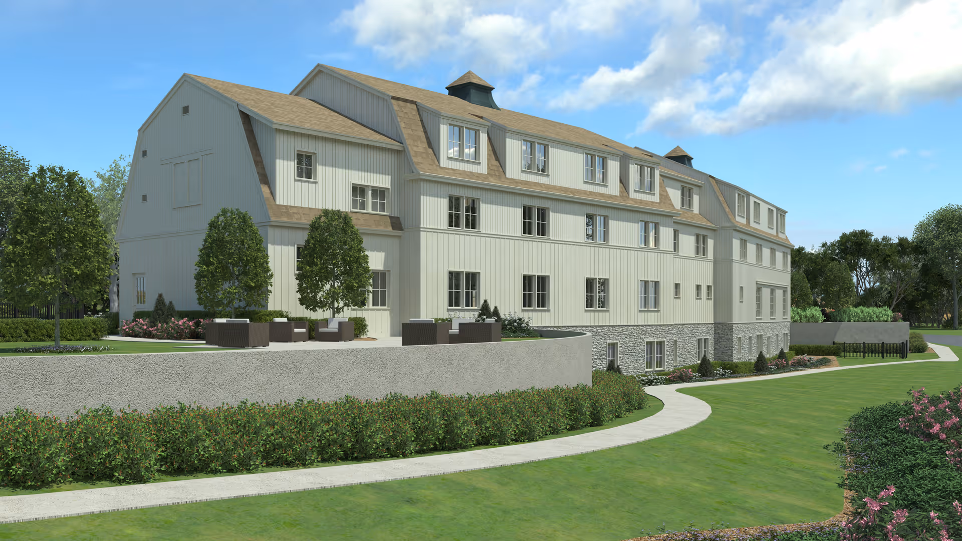 Exterior view of a large, multi-story assisted living facility building with white siding and a tan roof. The building is surrounded by well-maintained green lawns, shrubs, and trees under a partly cloudy blue sky. A curved concrete pathway runs along the front of the building.