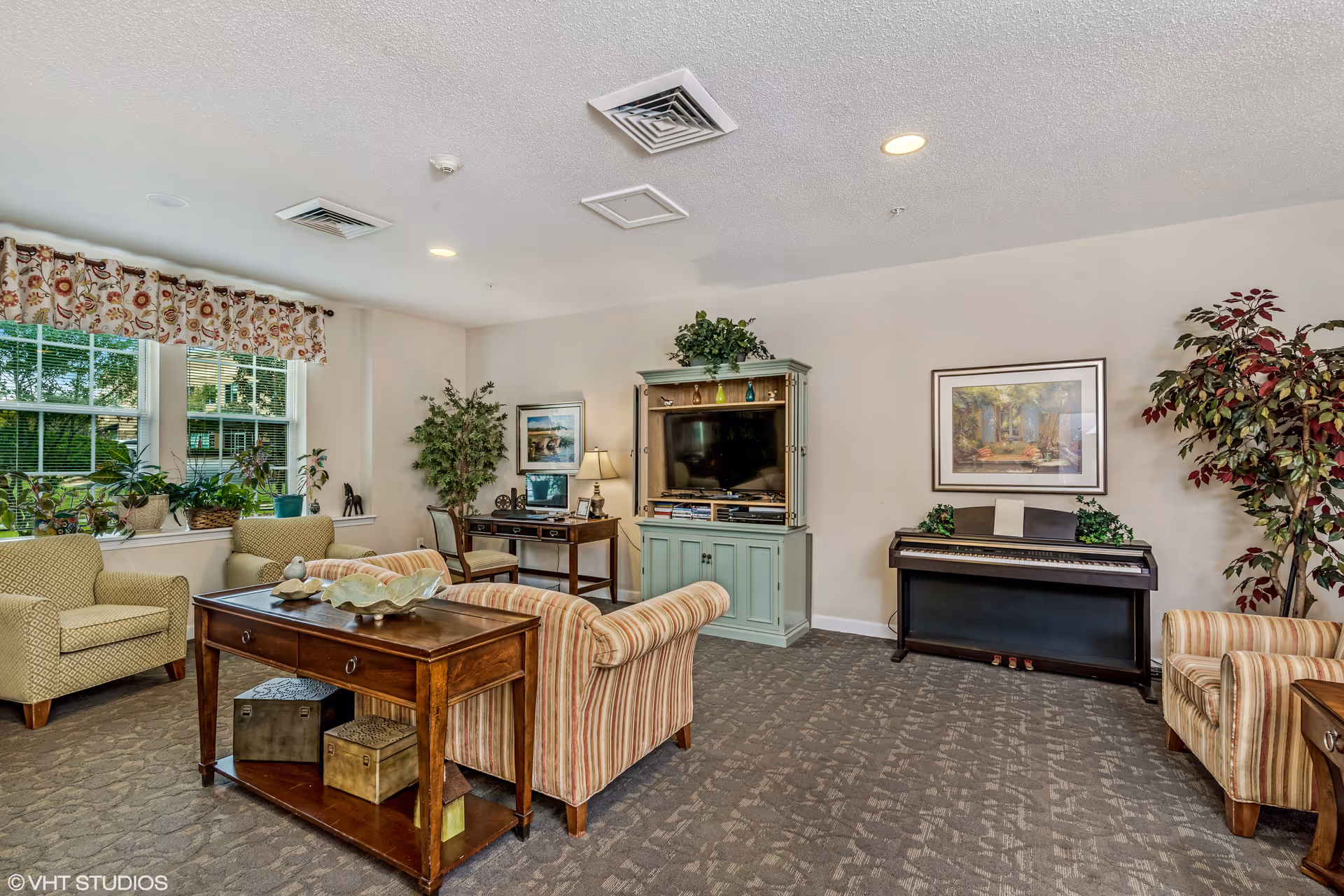 A cozy living room in a senior living facility with striped armchairs, a wooden coffee table, a green entertainment center with a TV, a piano, a desk with a chair, several potted plants, and framed artwork on the walls. Large windows with floral valances let in natural light.