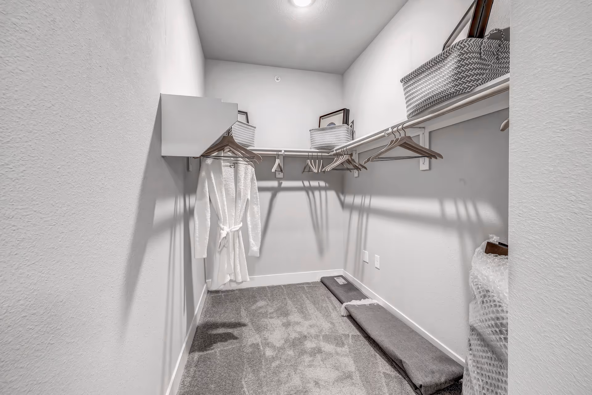 A walk-in closet with white walls and carpeted floor, featuring a white bathrobe hanging on a wooden hanger, several empty wooden hangers on a rod, and woven storage baskets placed on a shelf above the rod.