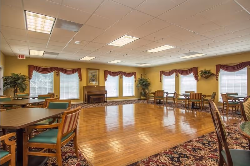 A spacious senior living community room with wooden flooring in the center and carpeted edges. The room has multiple tables and chairs arranged around the perimeter, large windows with red valances letting in natural light, a piano against the far wall, and potted plants in the corners. The ceiling has recessed lighting panels.
