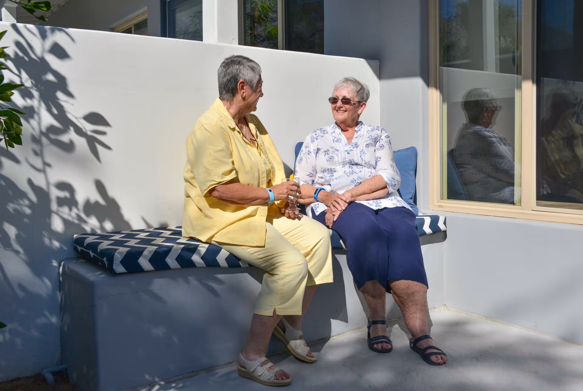 Two elderly women sitting and chatting on a cushioned bench outside a building with a window reflecting one of the women. One woman is wearing a yellow outfit and the other is wearing a white floral blouse with navy pants. They appear to be enjoying a sunny day.