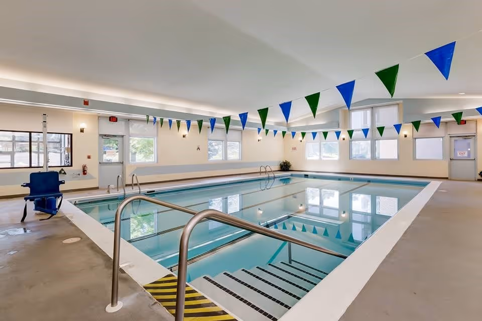 Indoor swimming pool area with steps and handrails, a blue pool lift chair, and blue-and-green pennant flags hanging from the ceiling.