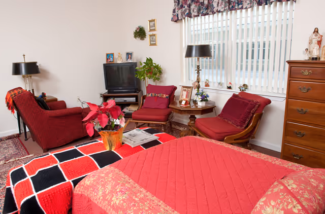 A cozy senior living room with a red and black checkered blanket on a bed in the foreground. The room features two red cushioned chairs, a wooden side table with a lamp and decorative items, a small TV on a stand, a wooden dresser, and a window with vertical blinds and floral valance. There are framed photos and plants decorating the space.