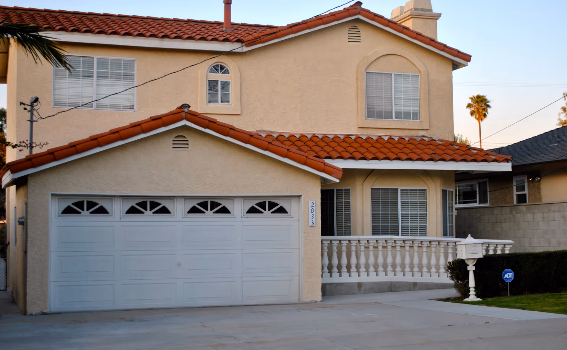 Exterior view of a two-story beige stucco house with a red tile roof, white garage door, and white balustrade on the front porch. There is a white mailbox and an ADT security sign on the lawn next to the driveway.