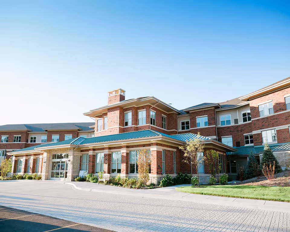 Exterior view of a senior living facility building with red brick walls, large windows, and a green roof under a clear blue sky. The entrance area is surrounded by small trees, shrubs, and benches along a paved driveway.