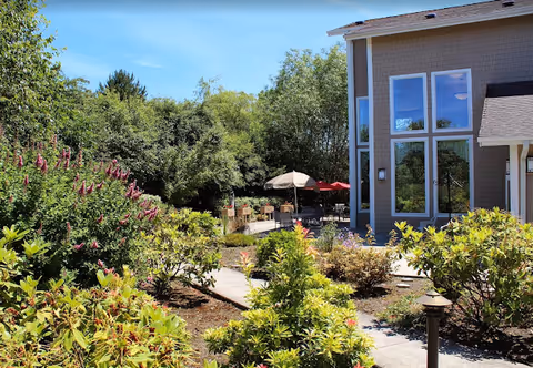 Outdoor garden area with lush green bushes and flowering plants along a paved walkway leading to a patio with tables, chairs, and umbrellas next to a building with large windows under a clear blue sky.