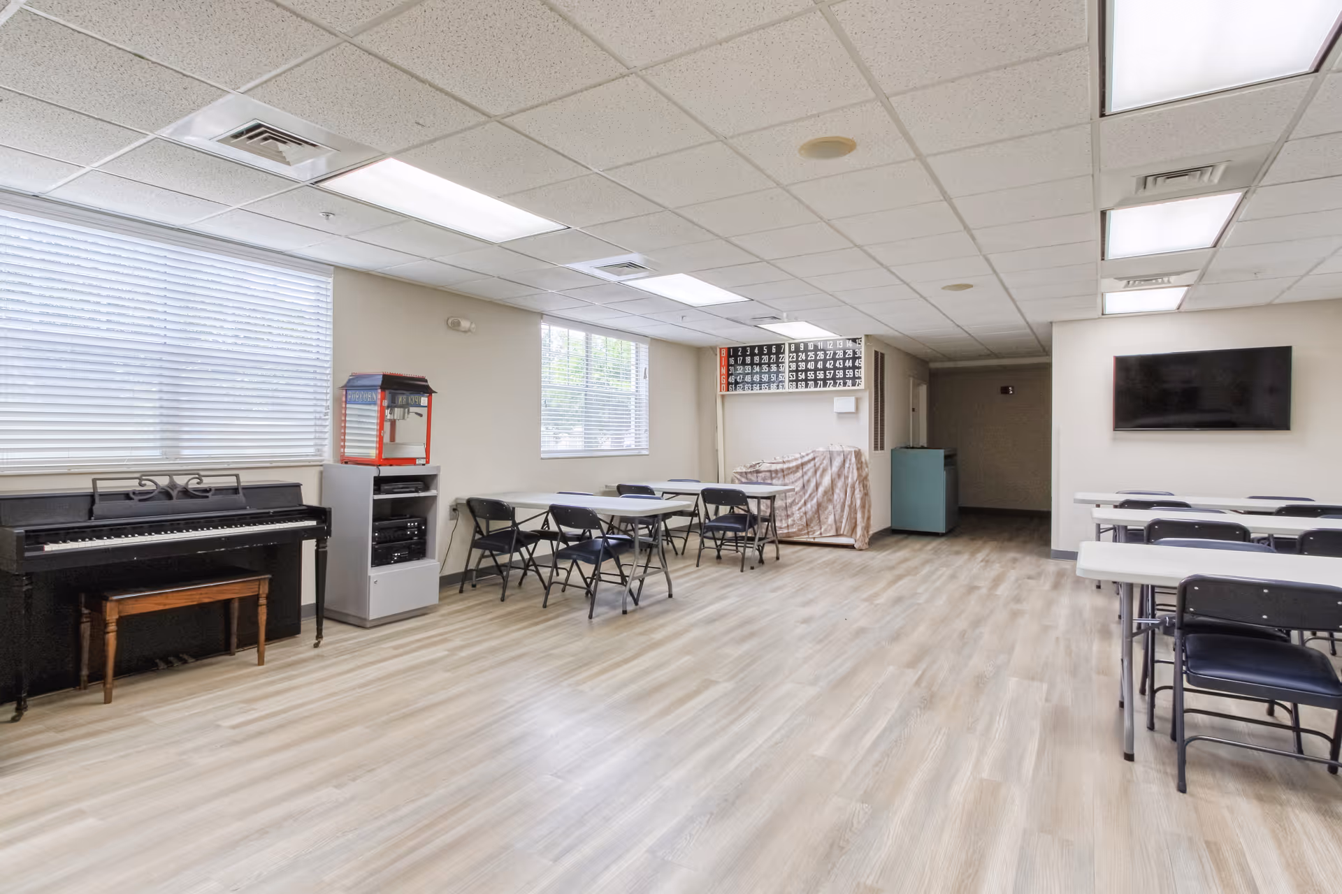 Large multipurpose activity room with folding tables and chairs, a piano, TV, and a popcorn machine.
