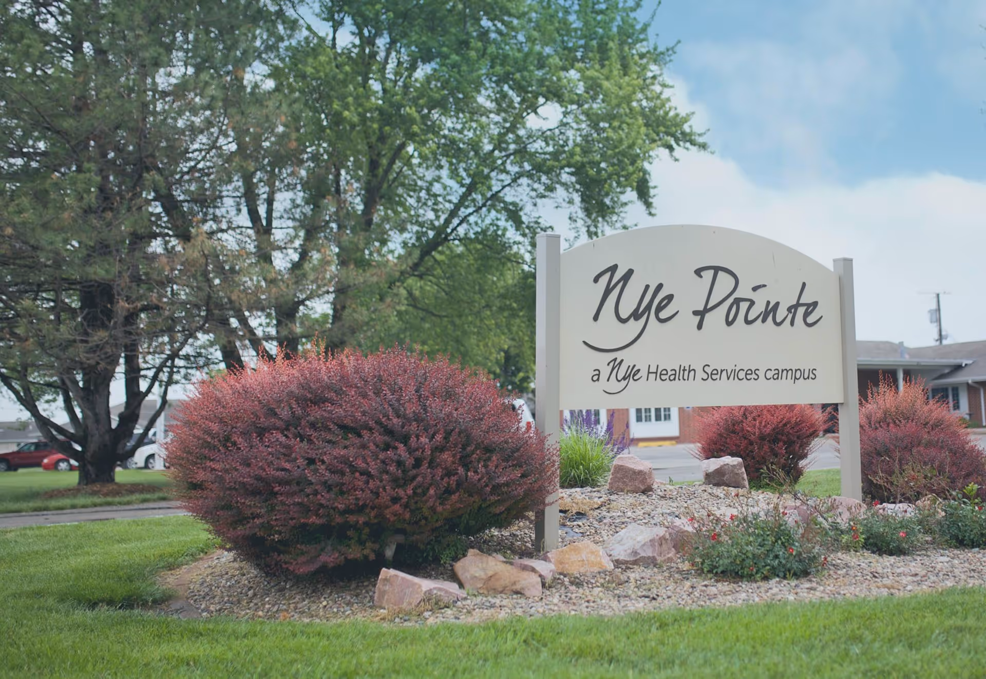 Outdoor view of a landscaped area with bushes, rocks, and grass in front of a sign that reads 'Nye Pointe a Nye Health Services campus' with trees and a building in the background under a partly cloudy sky.