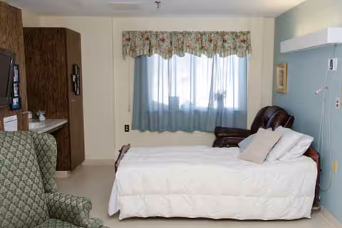 A senior living bedroom with a neatly made bed, armchair, desk, and window with curtains.