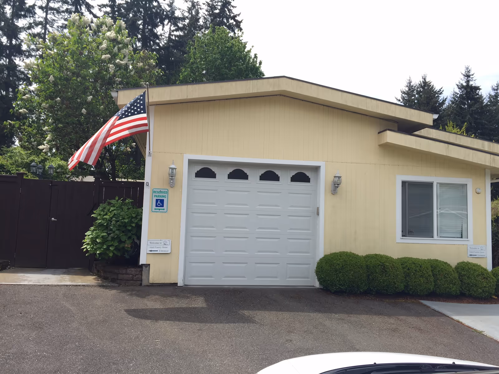 Front exterior of a small yellow single-story building with a closed white garage door, an American flag, and manicured bushes.