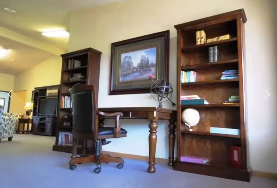 A cozy interior space featuring a wooden desk with a black office chair on wheels. On the desk is a decorative armillary sphere. Behind the desk, there is a framed painting of a serene outdoor scene. Flanking the desk are two tall wooden bookshelves filled with books and decorative items, including a globe. In the background, there is a glimpse of a living area with a patterned armchair and a TV stand with a television.