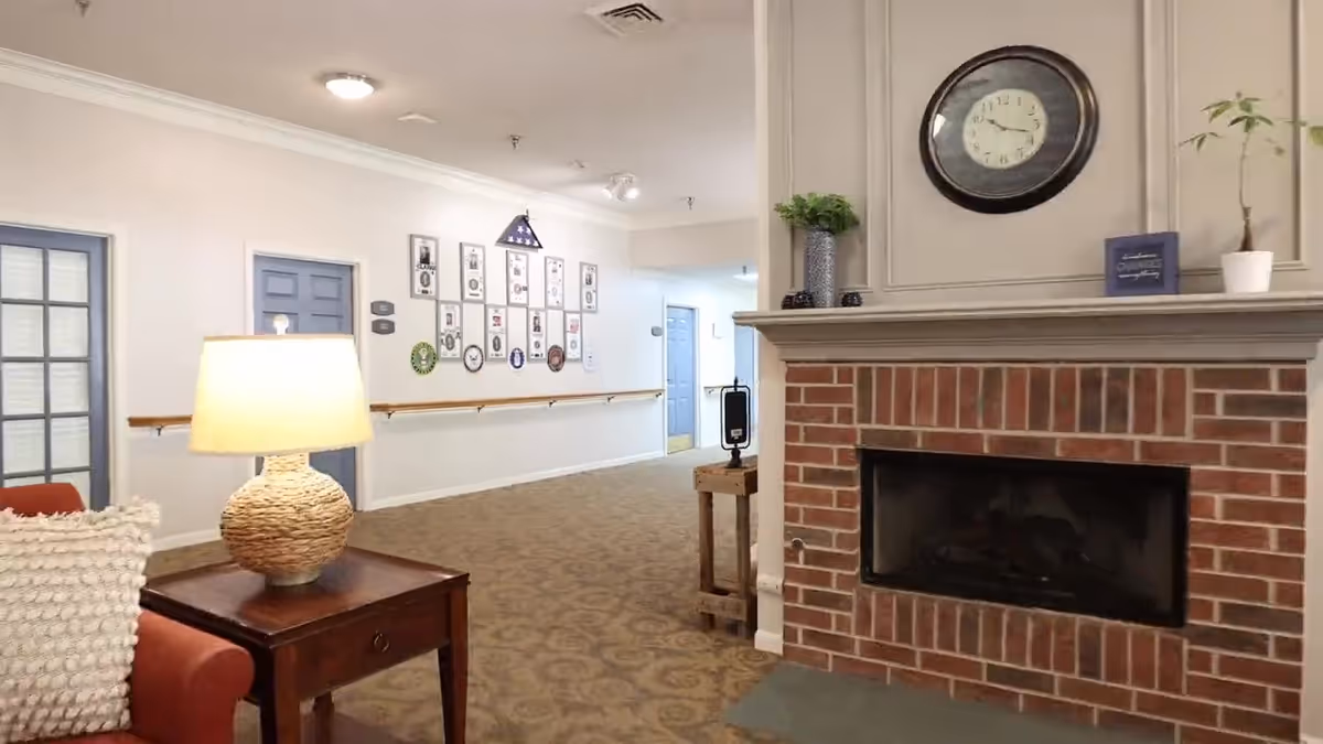 Assisted living common room with a brick fireplace and clock, a lamp on a side table beside seating, and a hallway with framed photos.