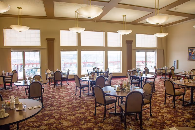 Spacious dining room with round tables and chairs set for meals, large windows and pendant lights.