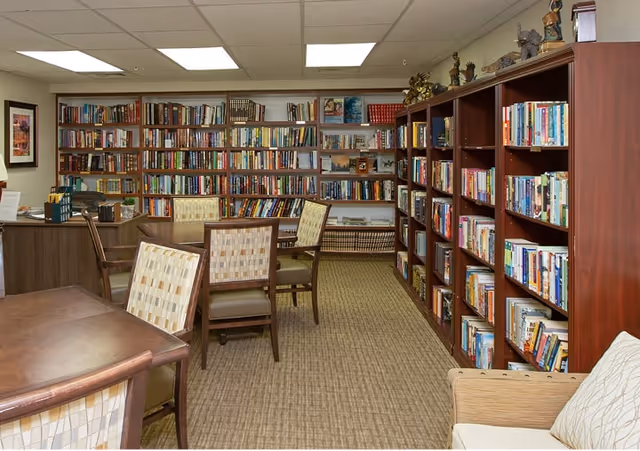 Interior view of a library room in a senior living facility with multiple wooden bookshelves filled with books, several chairs with patterned upholstery around tables, and a desk in the corner. The room has a carpeted floor and a drop ceiling with fluorescent lighting.