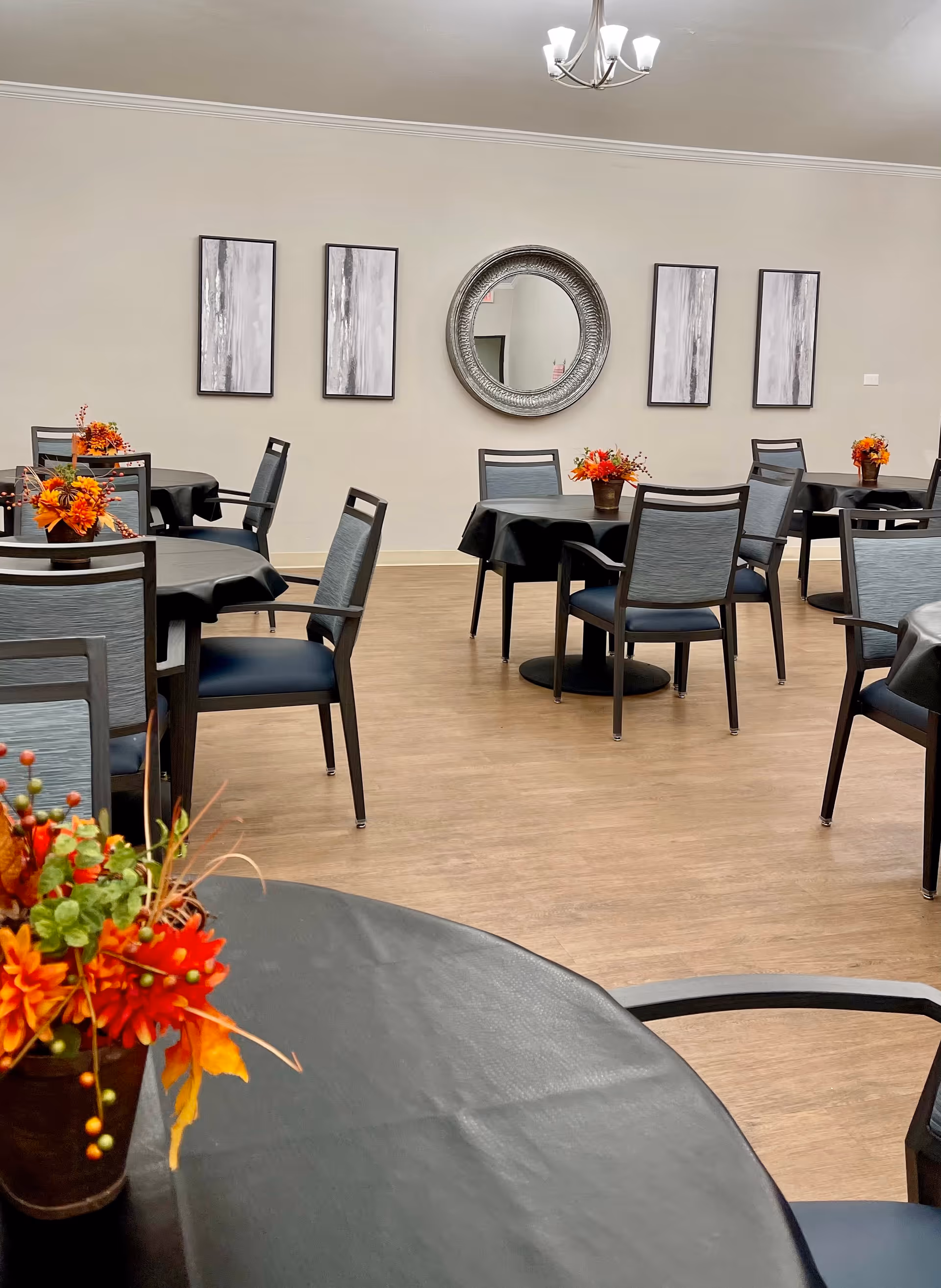 A dining room with round tables and chairs topped with autumn floral centerpieces and wall art on a neutral wall.