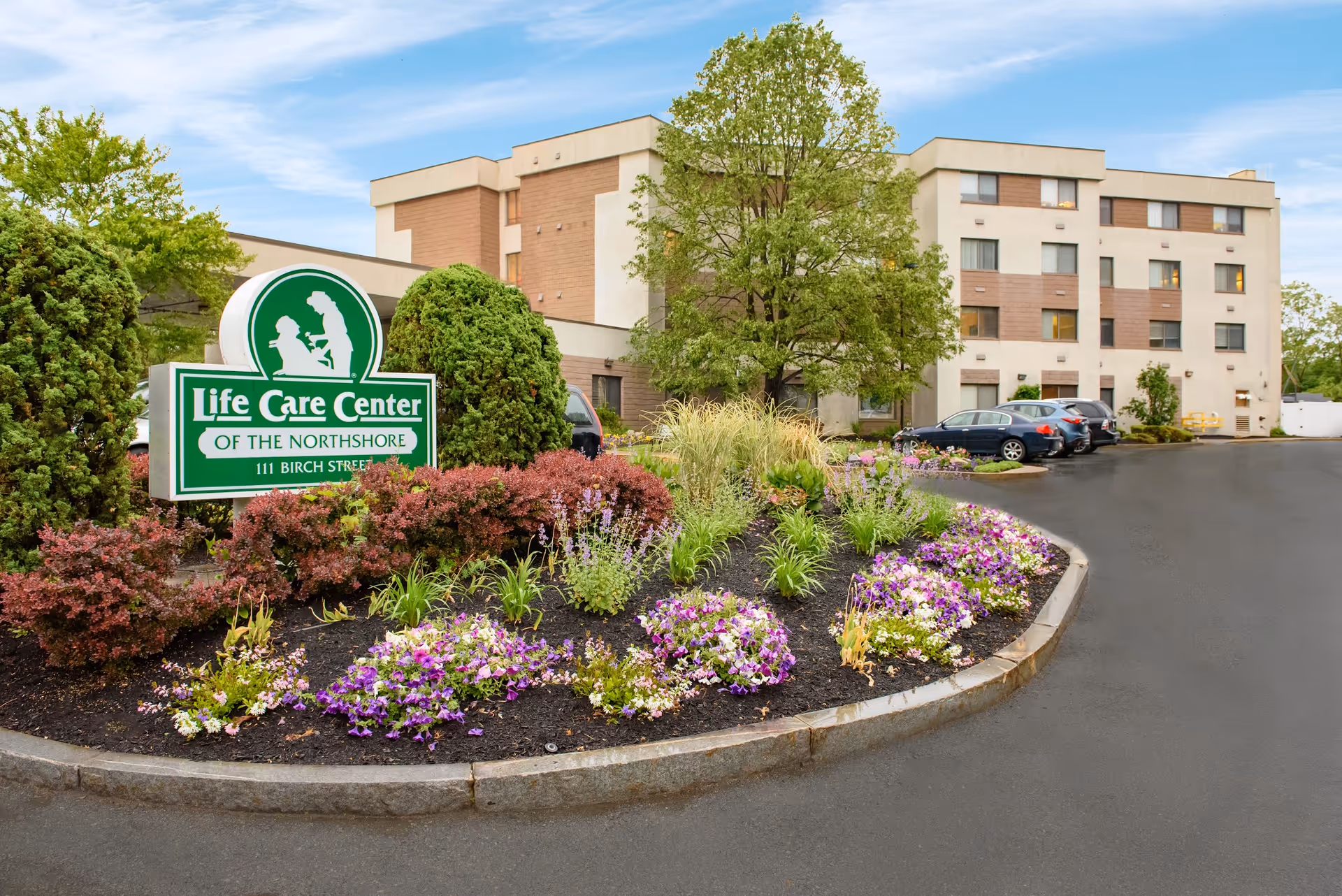 Front exterior of the Life Care Center of the Northshore showing the building, entrance sign, landscaped flower beds, and parked cars.