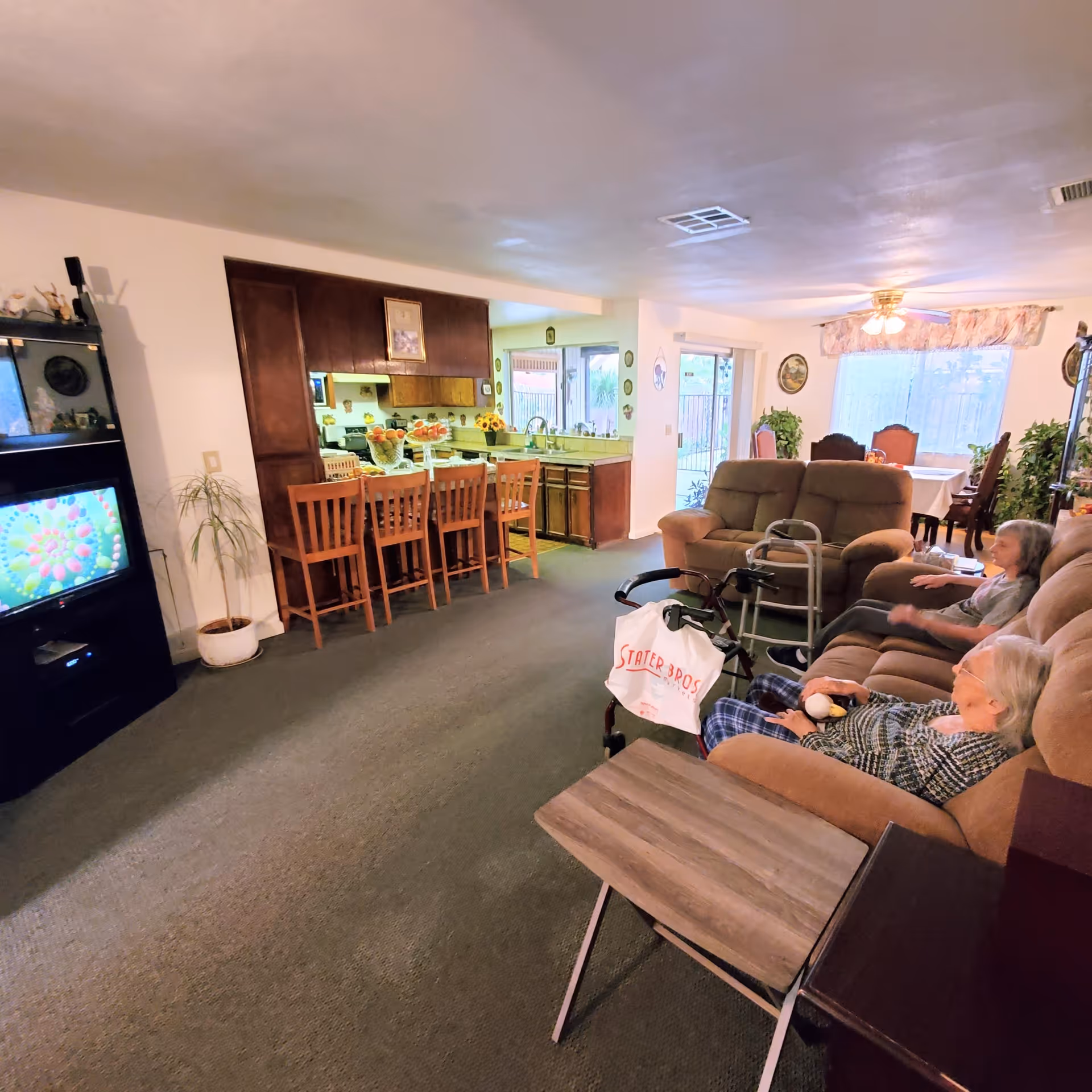 A cozy living room area in a senior care facility with two elderly women sitting on a brown couch. A walker with a Stater Bros. bag is nearby. The room includes a TV on a black stand, a wooden table, and a kitchen with a breakfast bar and wooden chairs in the background. The dining area is visible further back with a table and chairs near large windows with curtains.