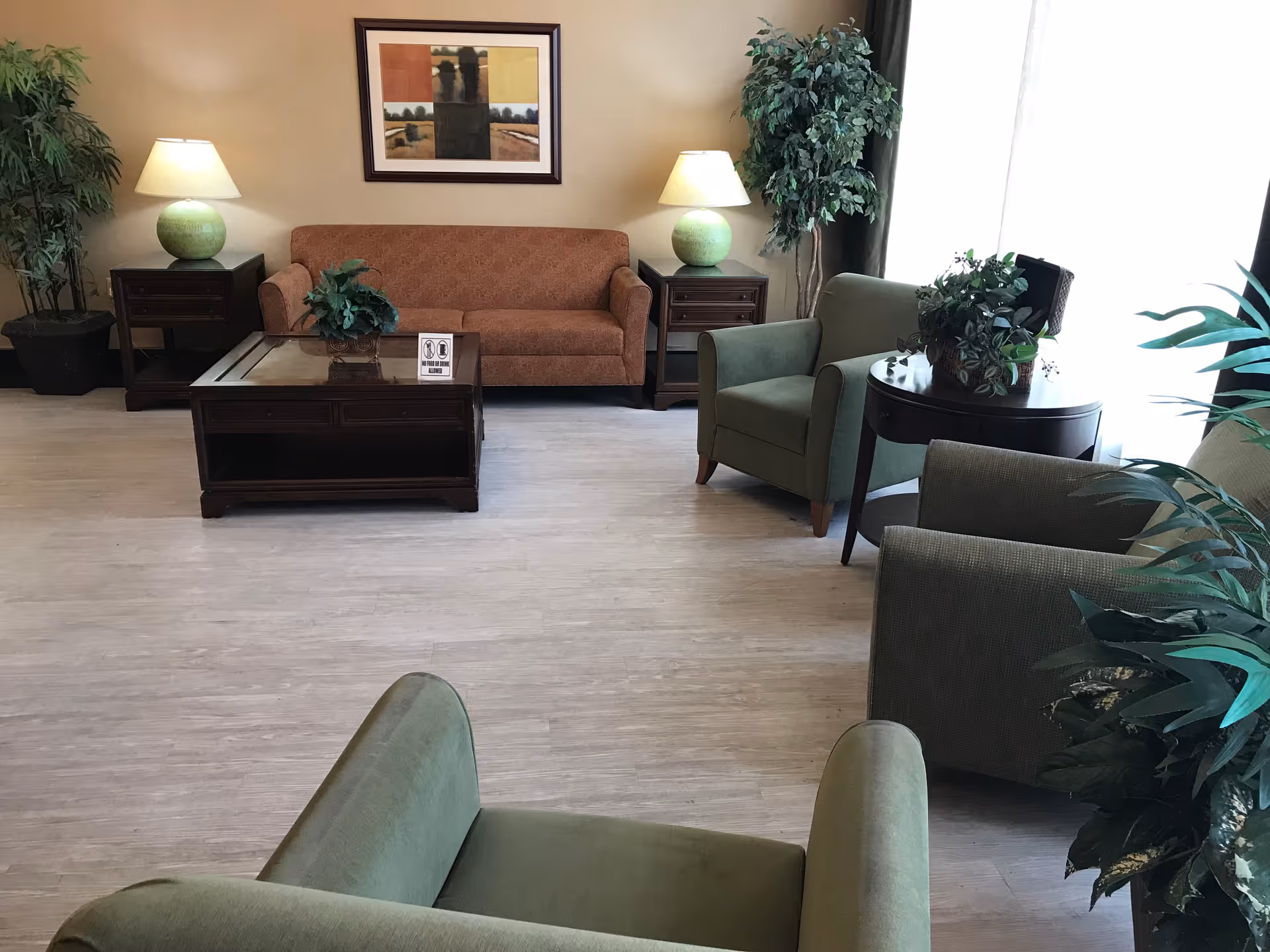 A well-lit sitting area with a brown sofa, several green armchairs, coffee tables, lamps, and potted plants.