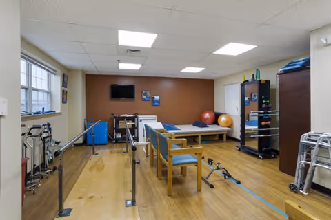 A physical therapy or rehabilitation room with wooden flooring, parallel bars for walking exercises, a treatment table with pillows, exercise balls, chairs, and various exercise equipment. The room has a brown accent wall with a mounted TV and framed pictures, and windows letting in natural light.