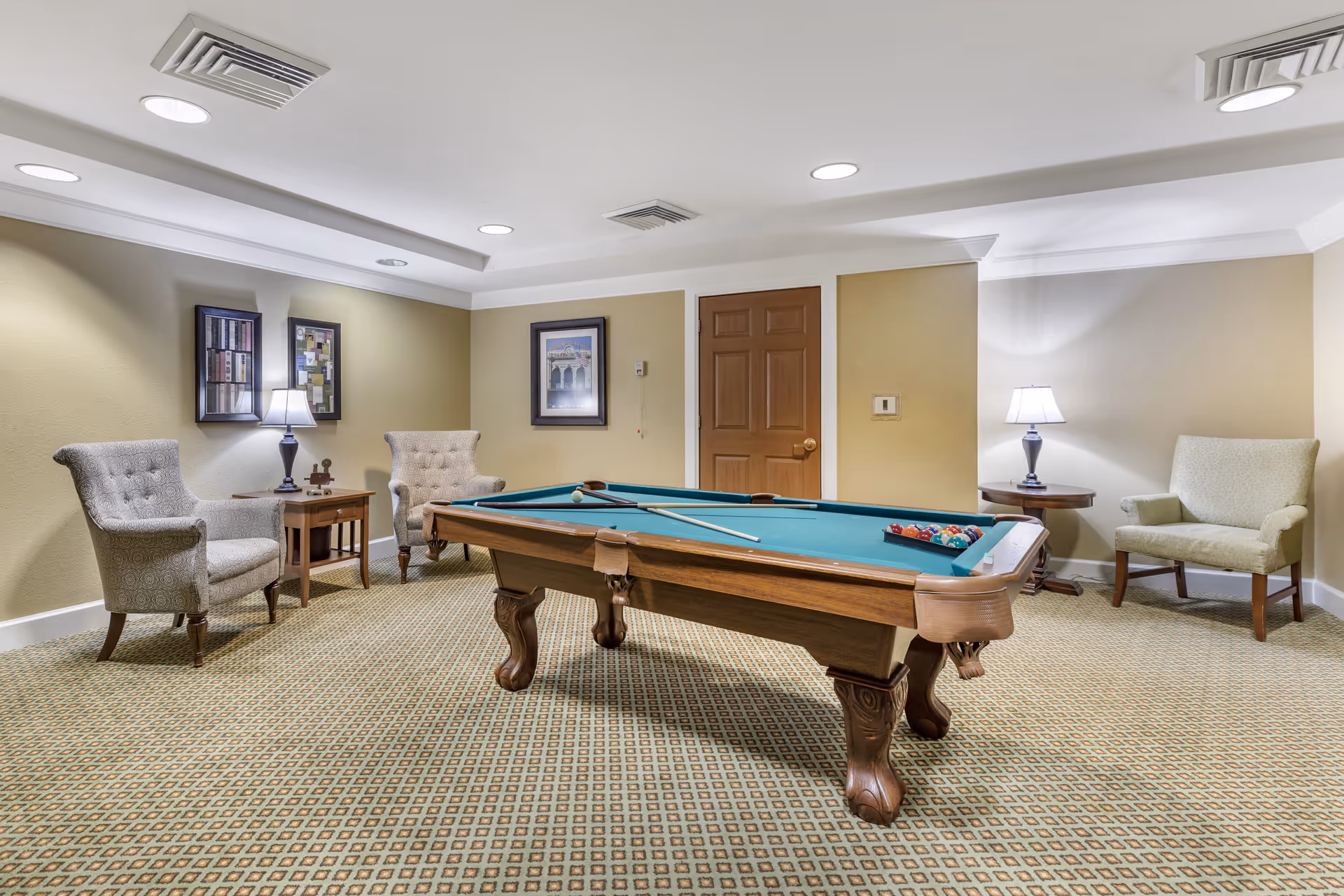 A well-lit room with a pool table in the center, surrounded by three upholstered armchairs and two side tables with lamps. The walls are beige with framed artwork, and there is a wooden door in the background. The carpet has a patterned design.