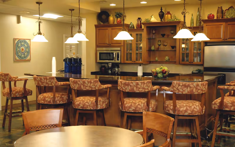 Interior view of a kitchen and dining area with wooden cabinets, a microwave, a refrigerator, and a kitchen island with six cushioned bar stools. Pendant lights hang above the island, and there is a round dining table with chairs in the foreground. Decorative items and plants are placed on top of the cabinets.